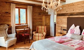 A cozy bedroom with wooden walls and a chandelier. There is a comfortable bed, an armchair, and a small table with chairs.