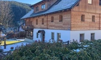 A picturesque wooden house with a snow-covered roof and green bushes in the foreground. In the background, gentle hills and a clear sky can be seen.