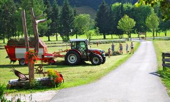 A tractor is working in a green field with a trailer. In the background, there are people busy with hay, and trees framing the landscape.
