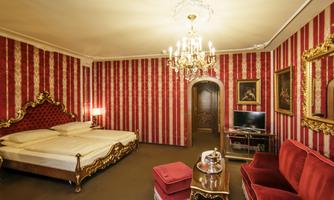 An elegant room with a red-striped background and opulent furnishings. The room features a large bed, a cozy seating area, and a crystal chandelier.