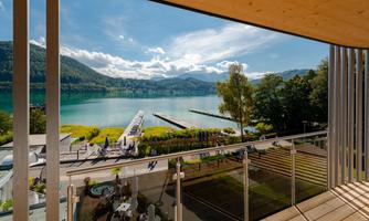 A beautiful view of a clear lake, surrounded by mountains and green trees. In the foreground is a modern terrace with plenty of natural light.