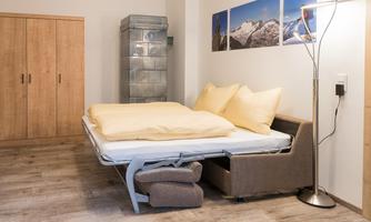 A simple bedroom with a fold-out bed and yellow pillows. In the background, light wooden wardrobes and pictures of mountains can be seen.