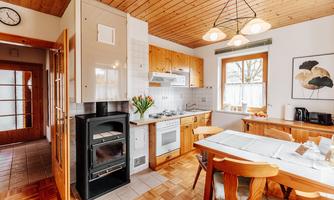 A modern kitchen with wooden cabinets and a cozy dining area. Fresh flowers are on the table next to the stove, and there are large windows for plenty of natural light.