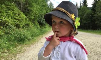 A little girl with a gray hat and a pink scarf stands on a gravel path. She gazes thoughtfully into the distance and has a finger on her lips.