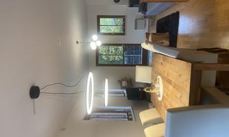 A modern dining room with a large wooden table and six white chairs. In the background, windows and a cozy living room can be seen.