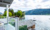 A view from a terrace of a calm lake with boat docks. In the background, gentle hills and a blue sky with clouds can be seen.