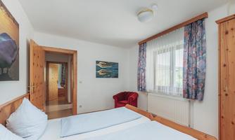 A cozy bedroom with two beds and a red armchair. There are windows with curtains and pictures on the walls.