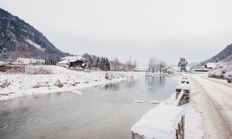 A wintry landscape with snow-covered mountains and a calm river. The surroundings are quiet and idyllic, with houses and trees along the bank.