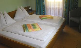 A cozy bedroom with a large double bed and colorful cushions. The room has light-colored walls and a window with curtains.