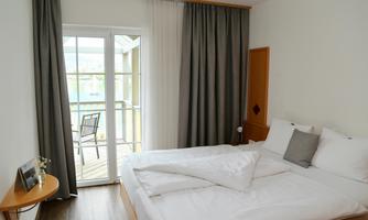 A bright bedroom with a large bed and a chair on the balcony. Large windows and modern curtains create a pleasant atmosphere.