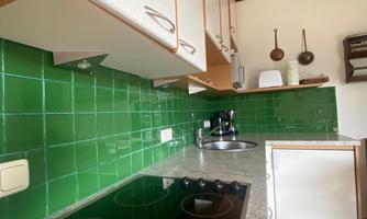 A modern kitchen with green tiles and wooden shelves. The countertop is made of grey stone and a stove is visible.