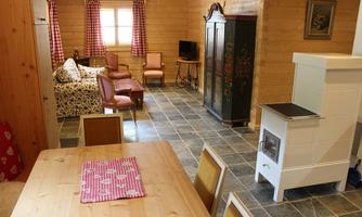 A cozy living room with a table, chairs, and a stove. In the background, there is a sofa and a television, surrounded by wooden paneling.