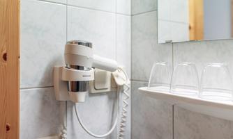 A hair dryer on the wall in a bathroom. Next to the hair dryer, several glasses are on a shelf.