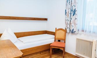 A simple room with a bed, a chair, and a table. The walls are light and a window provides daylight.