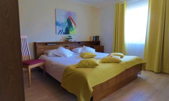 A cozy bedroom with a large bed and yellow pillows. The walls are bright and a window provides plenty of daylight.