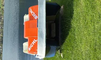 A pedal boat with orange seats is on green grass at the shore of a lake. In the background, there are trees and a calm water surface visible.