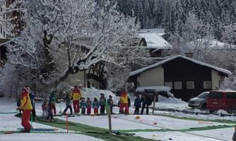 A group of ski instructors and children stands in a snowy meadow. In the background, there are snow-covered trees and cabins.