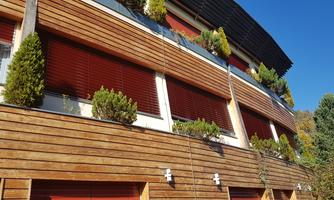 A modern building with a wooden facade and large window shutters. Plants and bushes can be seen on the balconies.