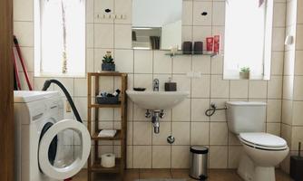 A modern bathroom with washing machine, sink, and toilet. White tiles and large windows create a bright atmosphere.