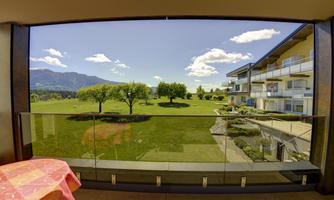 A beautiful view from a balcony overlooking a green meadow with trees and surrounding mountains. The sky is blue with a few white clouds.