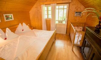 A cozy bedroom with wooden paneling, a large bed with decorative pillows, and a child's bed. Plenty of daylight streams in through the windows, and a flower adorns the table.