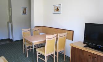 A simple living room with a table and four chairs. A TV is on the wall and some pictures are hanging.
