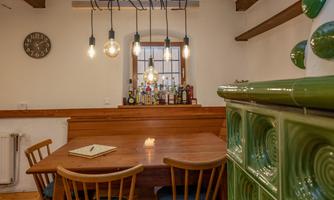 A cozy dining area with a wooden table and chairs. Modern lamps hang over the table, while a green tiled heater stands on the wall.