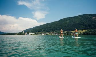 Two people are paddling on stand-up paddleboards on a calm lake. In the background, there are green hills and a sky with some clouds.