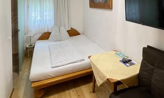 A cozy room with a double bed, a table, and a chair. There is a window with curtains and a television on the wall.