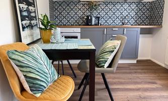 A modern kitchen corner with a table and two chairs. The walls are decorated with blue tiles, and there are plants as well as colorful cushions on the chairs.