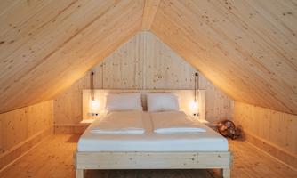 A cozy bedroom in the attic with wooden paneling and soft lighting. The bed is neat and inviting, perfect for a relaxing getaway.