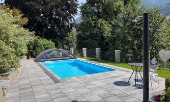 A beautiful swimming pool in a garden, surrounded by trees and plants. Next to the pool, there is a table with chairs for cozy hours outdoors.