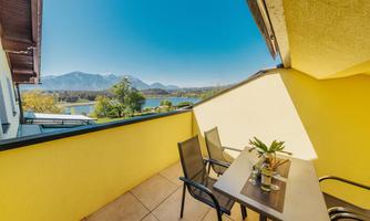 A cozy balcony with a table and chairs that offers a view of the mountains and a nearby lake. The sky is clear and sunny.