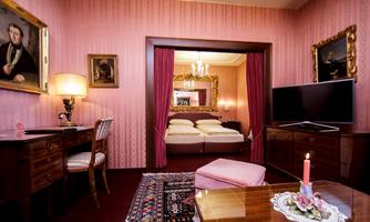 An elegant room with pink walls and classic furniture. In the background, a bedroom with two beds can be seen.