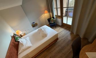 A cozy room with a bed, a table, and a chair. Large windows offer a view of a balcony.