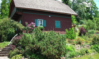 A traditional red wooden house surrounded by a blooming garden. The garden is full of colorful plants and bushes.
