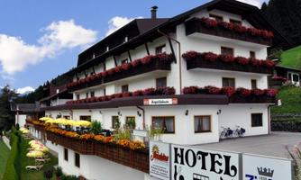 A cozy hotel with several floors and balconies adorned with flowers. The surroundings are green and mountainous, perfect for a relaxing stay.