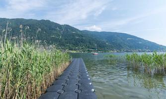 A walkway leads over the water to a beautiful lake, surrounded by green shore plants. In the background, gentle hills and blue sky can be seen.