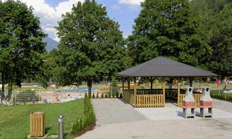 A beautiful park with a barbecue area and many trees. In the background, a swimming pool can be seen.