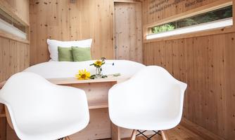 A cozy interior with wooden paneling and large windows. There is a round table and two modern chairs, decorated with flowers.