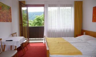 A simple room with a double bed and a table. Large windows offer a view of the green nature outside.