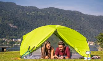 A couple relaxes in a green tent on a meadow. In the background, mountains and a lake can be seen.