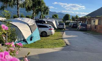 A picturesque campsite with tents and cars, surrounded by trees and blooming flowers. The sun shines on a tranquil landscape with mountains in the background.