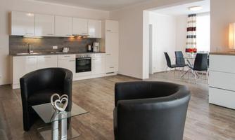 A modern living room with an open kitchen. The furnishings are clear and bright, featuring comfortable chairs and a dining area.