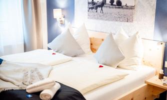 A cozy bedroom with a double bed and large, white pillows. On the wall hangs a picture of riders, and there are decorative elements like towels and small red hearts on the pillows.