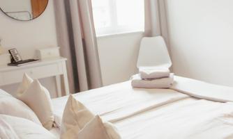 A bright bedroom with a large bed and cozy pillows. The room has white walls and a window that lets in plenty of light.
