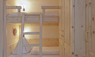 A cozy room with a loft bed made of light wood. A small light hangs on the wall, and there is a wooden wardrobe.