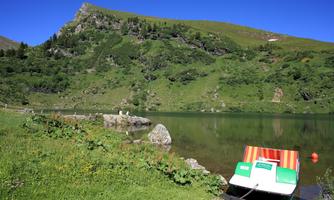 A quiet lake surrounded by green hills and trees. A boat lies at the shore and reflects in the water.