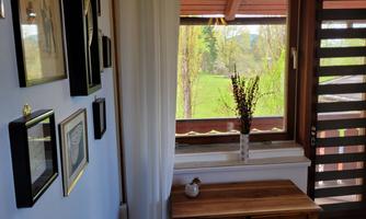 A cozy interior with a wooden table and framed pictures on the wall. Through the window, one can see greenery and light streams in.