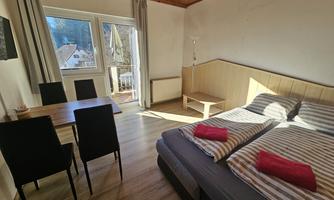 A cozy bedroom with two beds, striped bedding, and red towels. A small table and chairs complement the decor, and large windows provide daylight.